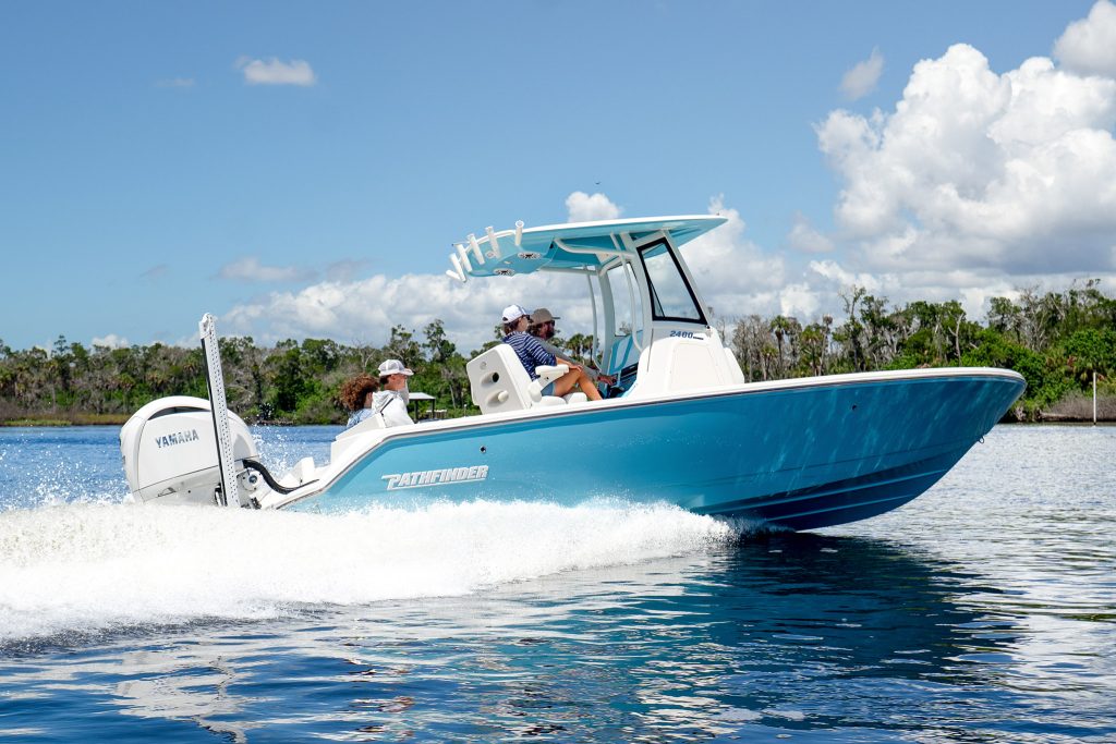 Teal and white Pathfinder 2400 Hybrid running at speed on blue water with two people aboard, a wooded shoreline and dock visible in the background under a partly cloudy sky.