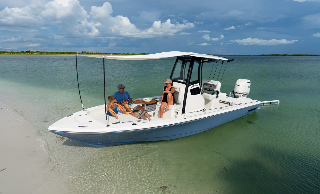 Pathfinder 2600 Hybrid beached near a sandy shoreline with its bow shade canopy extended, a family of three relaxing aboard under dramatic clouds.