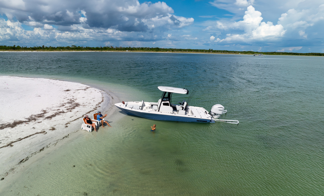 Aerial view of a Pathfinder 2600 Hybrid beached on a white sandbar under a partly cloudy sky, with people wading and relaxing on the sand beside it.