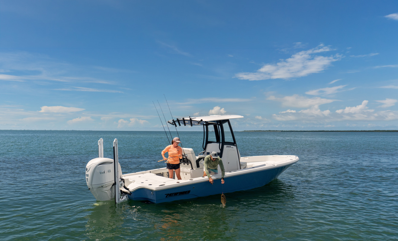 Two anglers aboard a blue and white Pathfinder 2600 Hybrid, one reaching over the gunwale while fishing on calm open water.