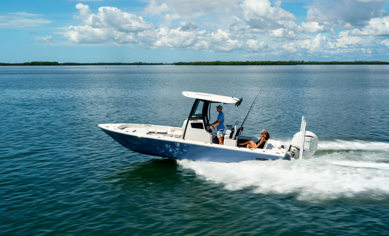 Pathfinder 2600 Hybrid running at speed across blue water with a wooded shoreline in the distance, two people aboard with fishing rods visible.