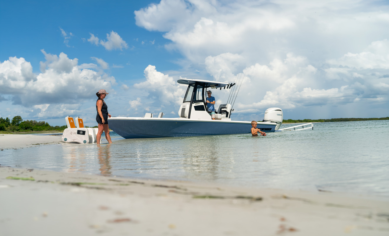 Low-angle, water-level view of a family beside a Pathfinder 2600 Hybrid beached on a sandbar, with a cooler, a man at the helm, and a child near the swim platform.