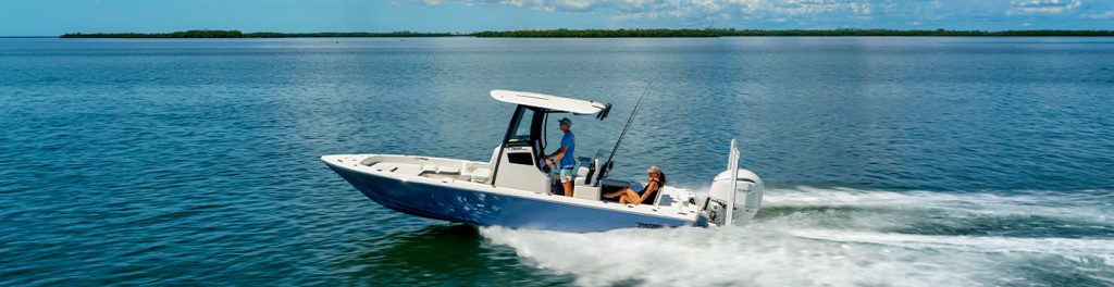 Pathfinder 2600 Hybrid running at speed across blue water with a wooded shoreline in the distance, two people aboard with fishing rods visible.